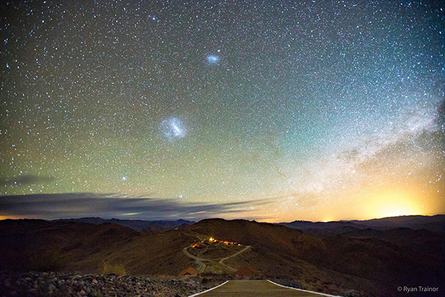An astrophoto of the Magellanic Clouds, which look like two wispy clouds with bright centers - along with thousands of stars - above Las Campanas Observatory in the desert of Chile